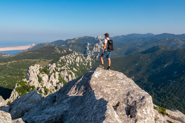 Fototapeta premium A lone hiker stands victoriously on a dramatic rocky peak in the Velebit mountains, overlooking the stunning Adriatic Sea coast in Kvarner Bay, Croatia on a summer day.