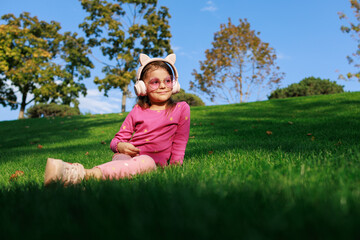 A girl wearing headphones and glasses is lying on the grass. cheerful child is playing and laughing in the park.