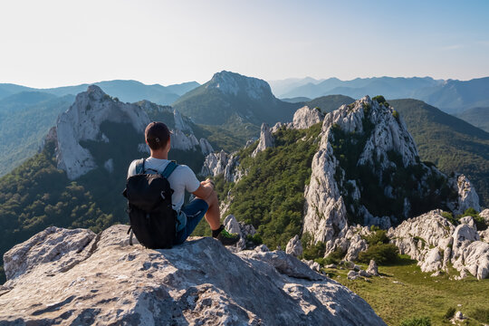 A male hiker with his backpack sits on a rocky peak in Croatia's Velebit mountains, contemplating the stunning and serene panorama of the majestic Dinaric Alps.