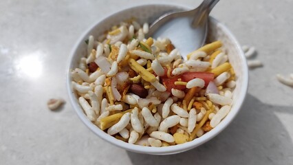 A closeup image of Indian street food bhel puri served in a white bowl with a steel spoon placed on a table with selective focus.