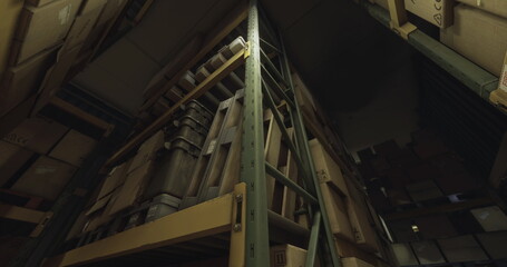 Fototapeta premium Tall shelves stacked with boxes fill a dimly lit storage facility. The structures create a maze like appearance, while shadows loom in the corners of the room.