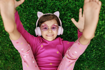 A girl wearing headphones and glasses is lying on the grass. cheerful child is playing and laughing in the park.