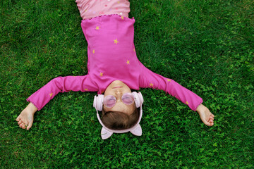 A girl wearing headphones and glasses is lying on the grass. cheerful child is playing and laughing in the park.