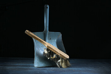 Old hand brush with wooden handle and a lot of dirt leaning on an aged metal dustpan against a dark background, still life of vintage cleaning tools, copy space
