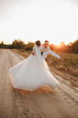 Groom twirls the bride on a sandy road at sunset. Wedding photo shoot in nature capturing motion, joy, and tenderness. Romantic atmosphere with warm evening sunlight and true love.