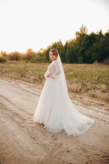 Bride in elegant white wedding dress standing on rural road at sunset. Romantic bridal portrait with soft golden light and natural outdoor background, symbolizing love, beauty, and serenity.