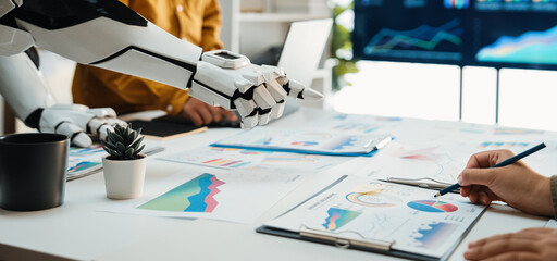 A human hand and a robotic arm collaborate on data analysis in a modern office space, showcasing innovative technology and teamwork through digital charts and graphs.Noogenesis
