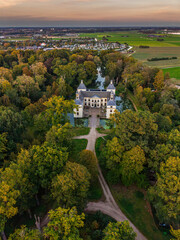 Aerial view of historic castle symmetrical twin towers and central hall framed by forest, moats, and tree-lined path, with open fields and residential zone beyond under warm sunrise sky.