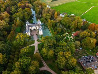 Aerial view of historic castle symmetrical twin towers and central hall framed by forest, moats, and tree lined path, with open fields and residential zone beyond under warm sunrise sky.