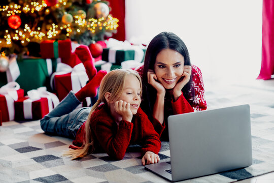 Mother and daughter enjoying Christmas together, using a laptop near a festive tree surrounded by presents, sharing joyful moments