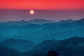 Sun descending over rolling hills, creating a tranquil atmosphere with a red sky and atmospheric haze