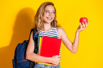 Young female student holding a red notebook and an apple in a casual outfit against a bright yellow...