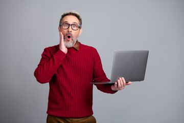 Surprised mature man in a red sweater holding a laptop against a gray background, expressing curiosity and interest