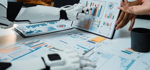 A robotic arm assists a business team in analyzing data charts and graphs in a modern office environment, showcasing collaboration and innovative technology integration.Noogenesis