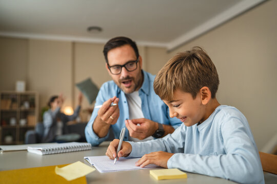 Father helping son doing homework with mother juggling in background
