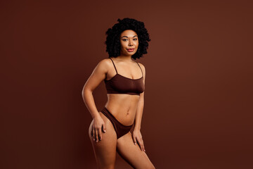 Naklejka premium Charming young woman in activewear posing against a solid brown background, symbolizing self-acceptance