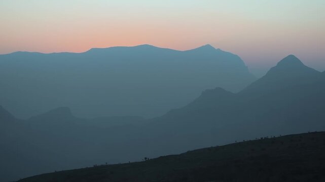 A serene and  view of the Al Hajar Mountains in Jebel shams, Oman at sunrise, with multiple layers of mountain peaks fading into the distance in a soft blue and pink haze. The first rays of morning li