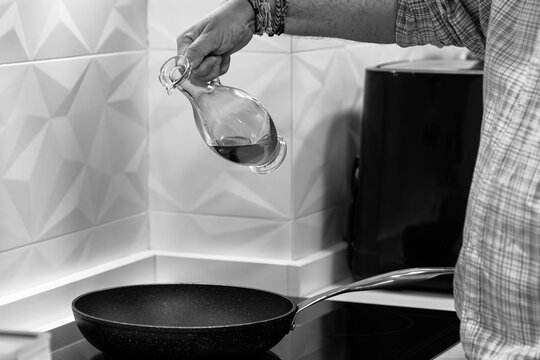 A man is pouring olive oil into a frying pan. The oil is in a glass jar. He is about to cook in the kitchen of his house. The frying pan is on a ceramic hob already turned on.