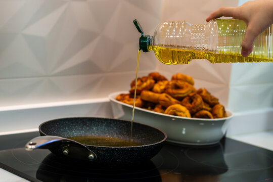 A person is pouring olive oil into the pan from a plastic bottle. He is in the kitchen. The pan is on the stove. In the background you can see a cooking dish filled to the top with a pile of freshly