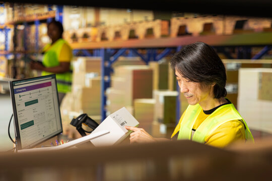 Asian logistics worker at a packing station manages order data entry, shipping labels numbers verification for inventory. Fulfillment center infrastructure supports online shopping.