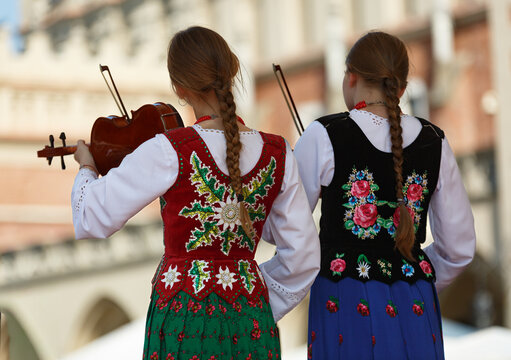 Two young girls in traditional folklore costiums embroidered with unique traditional pattern stand on their back playing violins