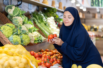 Smiling interested young european girl wearing islamic dress and chador selecting ripe red tomatoes while shopping in fruits and vegetables section of supermarket