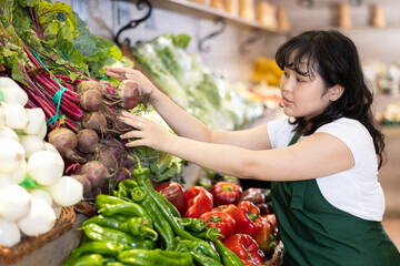 Skilled interested young asian girl in green apron working at farmers market, arranging fresh beets on vegetable display