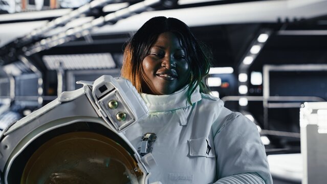 Portrait of jolly female astronaut holding helmet inside space station after completing surface mission on unfamiliar planet. Smiling african american cosmonaut preparing for return to Earth