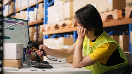 Sleepy asian staff member dealing with burnout in industrial warehouse, struggling to keep awake and ensure seamless package tracking service on software. Tired overworked employee. Camera A.