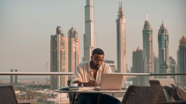 A young African man with short hair works on a laptop at a balcony overlooking a modern city skyline with tall buildings.