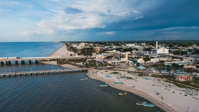 Aerial panoramic view of a coastal city with a pier, sandy beach, fishing boats, and amusement rides near the shoreline. The scene shows the mix of urban life, tourism, and maritime activity against t