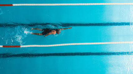 Aerial top view of a swimmer in a blue pool performing freestyle stroke between lane dividers. The image captures the dynamic movement of water and the athletic effort of competitive swimming.