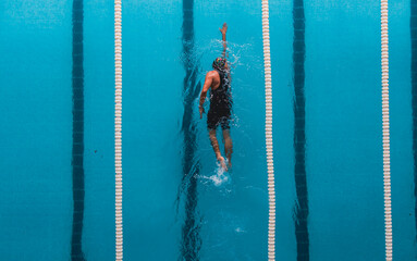 Aerial top view of a swimmer in a blue pool performing freestyle stroke between lane dividers. The image captures the dynamic movement of water and the athletic effort of competitive swimming.