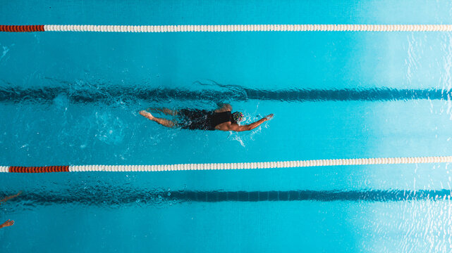 Aerial top view of a swimmer in a blue pool performing freestyle stroke between lane dividers. The image captures the dynamic movement of water and the athletic effort of competitive swimming.