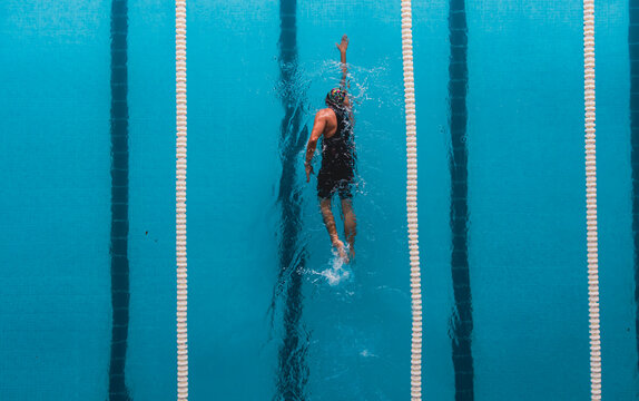 Aerial top view of a swimmer in a blue pool performing freestyle stroke between lane dividers. The image captures the dynamic movement of water and the athletic effort of competitive swimming.