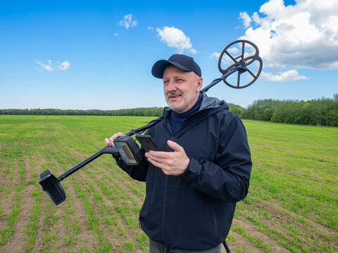 Man holding metal detector and smartphone in green field, checking coordinates or map for treasure hunting. Metal Detecting, GPS Navigation, Exploration, Outdoor, Travel.