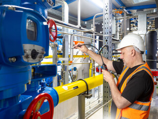 Technician in hard hat and hi-vis vest checking pipelines with a gas leak detector at a...