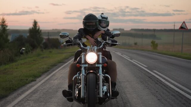 Two friends wearing helmets ride motorcycle on open road at sunset, passenger hugging rider from behind while headlight shines bright, capturing friendship and freedom of countryside travel at dusk