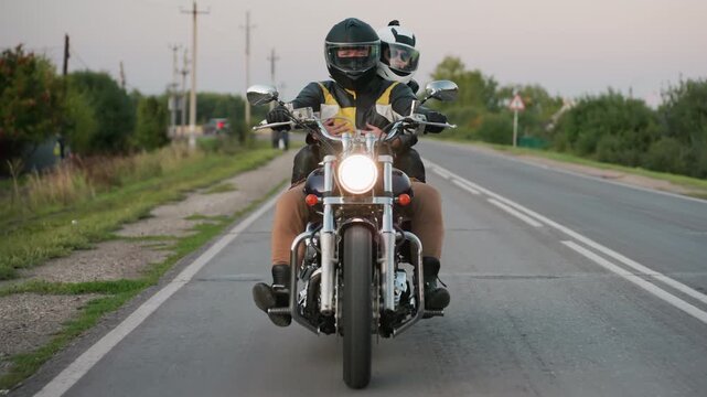 Motorcycle rider with passenger wearing helmets travels down countryside road at dusk, glowing headlight shining on asphalt as trees and roadside vegetation line horizon and sharp bend signpost