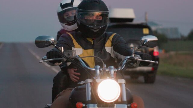 Close up of couple riding motorcycle on open road during evening, both wearing helmets and protective jackets, with soft sunset glow reflecting in visor