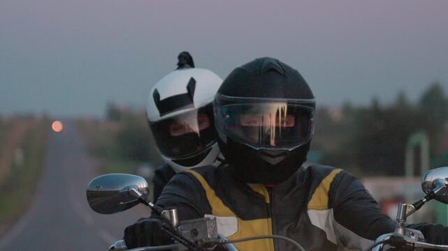 Close up of couple wearing protective helmets riding motorcycle uphill on countryside road during twilight, hands on handlebar, headlight glowing, relaxed mood, open air, freedom, long road ahead