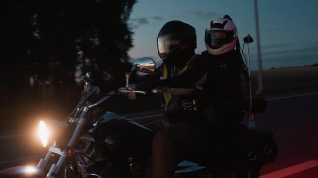 Motorcycle couple wearing helmets riding together on evening highway, bright headlight shining as they enjoy road trip at dusk, filmed from passing car with scenic countryside