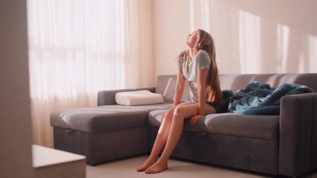 Banker seated on couch in bright room happily wiggles leg with joyful smile enjoying holiday morning while sunlight streams through window creating cheerful atmosphere with blanket beside her