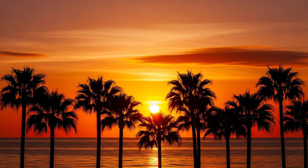 Obraz premium Palm trees silhouetted against a vibrant orange sunset over the ocean, creating a picturesque tropical scene with a warm and inviting atmosphere