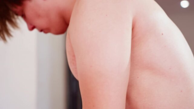 Guy with bare shoulder and upper back visible in gym setting, as he wears another shirt , showing skin texture and body detail under soft lighting, background blurred with punching bag