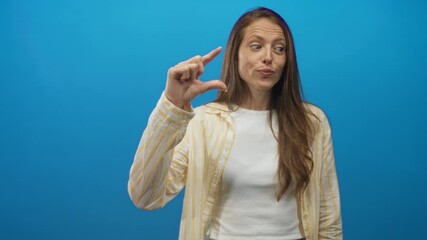 Woman with long hair pinching her fingers to show small size in a bright blue studio wearing a white tee and yellow striped shirt; playful skepticism.