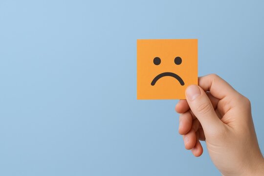 Sad face symbol displayed on orange sticky note held by hand against a blue background
