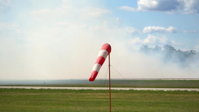Red and white striped windsock mounted on a tall pole at an airfield with heavy smoke on the background. The windsock is extended, showing the wind direction and strength