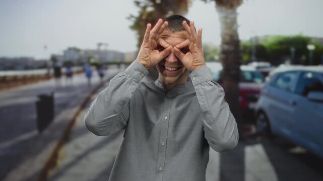 Man making binoculars gesture outdoors with a playful expression on a sunny street lined with palm trees and parked cars in the background.