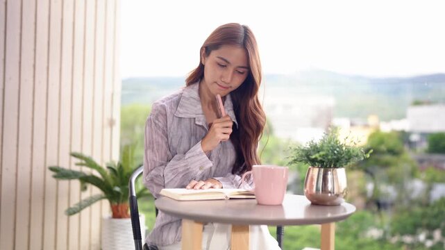 Asian woman writing journal pondering ideas on balcony home outdoor space for creative reflection and personal planning